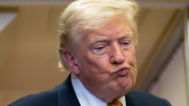A man with blonde hair, wearing a dark suit and red tie, purses his lips while looking downward against a plain tan background.