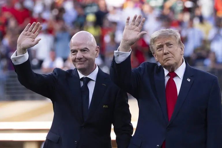 Two men in dark suits with ties wave to a crowd in stadium seating, one wearing a "FIFA" badge.