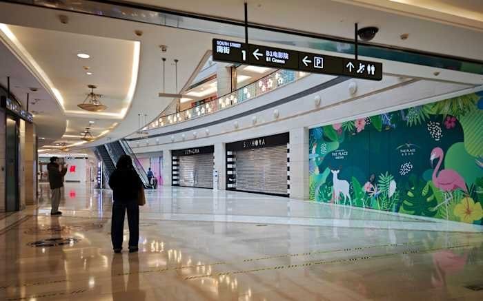 A nearly empty shopping mall corridor features two people standing near closed storefronts with lowered metal shutters, alongside a tropical-themed mural and overhead directional signage.