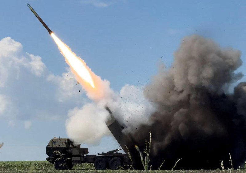 A military truck-mounted rocket launcher fires a rocket into the sky, producing a large plume of smoke and fire over a grassy field.