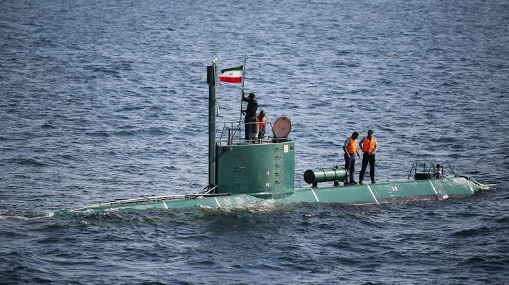 A green submarine surfaces in open water, with crew members in orange life vests standing on its deck beneath a flag with green, white, and red stripes.