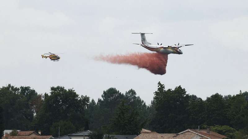 A white four-engine airplane marked "76" releases a large plume of red fire retardant above a tree-lined area, with a yellow and white helicopter flying nearby.