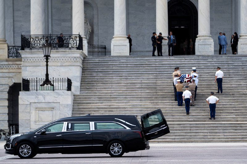A military honor guard carries a flag-draped casket down the steps of a columned building toward an open black hearse.