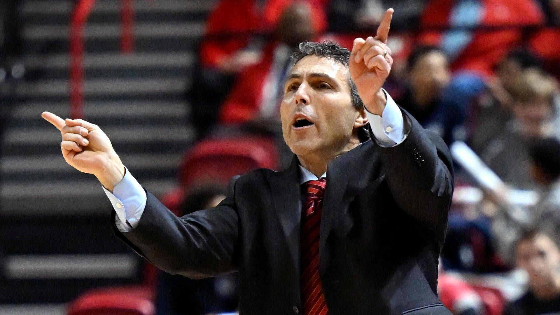 A man in a dark suit and red striped tie gestures with both index fingers pointing upward while shouting, set against a blurred background of red arena seating and spectators.